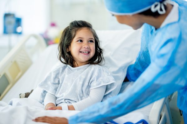 A sweet little girl of Hispanic decent sits up in her hospital bed as she talks with the doctor before her surgery.  She is wearing a hospital gown and is smiling as the doctor tries to put her at ease before wheeling her into surgery.